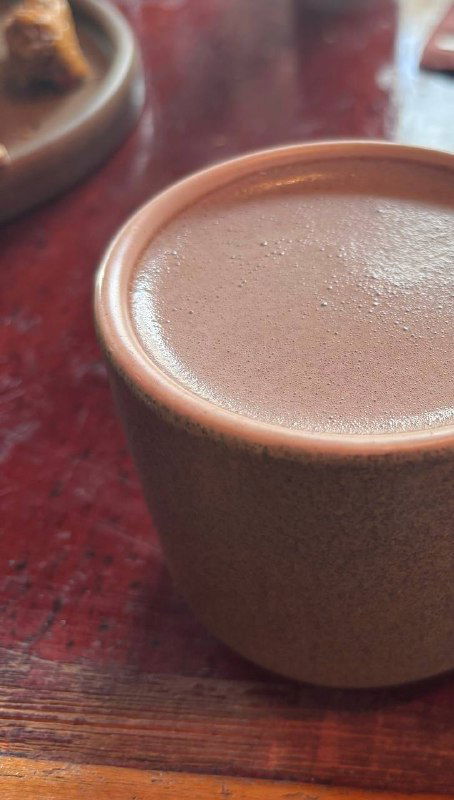 Крупный план чашки с тёплым какао на кокосовом молоке: кремовая поверхность напитка в простом керамическом стаканчике на столе.