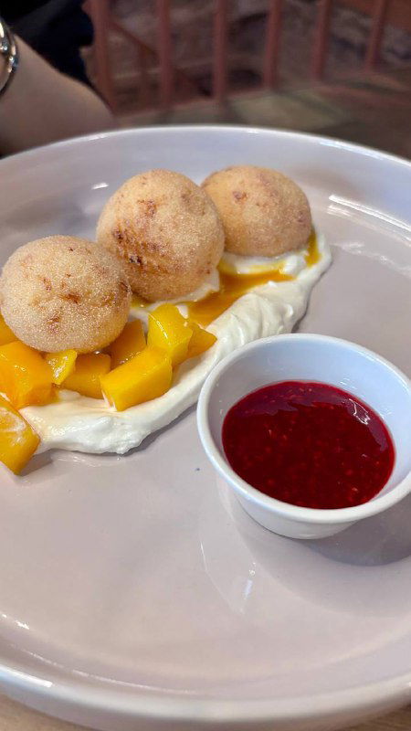 Десерт: шарики мороженого или профитроли на кремовой подложке рядом с кусочками манго и ягодным соусом в маленькой пиале на тарелке.