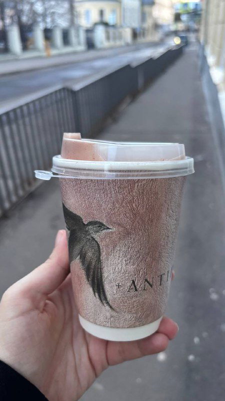 Рука держит бумажный стаканчик с логотипом кофейни на фоне городской улицы — уличный вид и крышка стакана крупным планом.