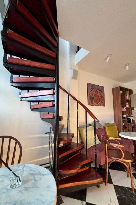 Спиральная деревянная лестница внутри кофейни — вид снизу, ступени и перила образуют виток, интерьер с картинами на стене.