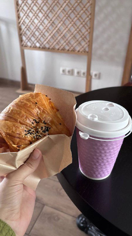 Круассан и бумажный стакан с горячим напитком на столе в кофейне при религиозном здании — завтрак в движении, крупный план.