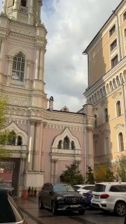 Двор и фасады зданий вокруг храма: пастельные тона стен, припаркованные машины на переднем плане и весенние растения у входа, городской пейзаж.