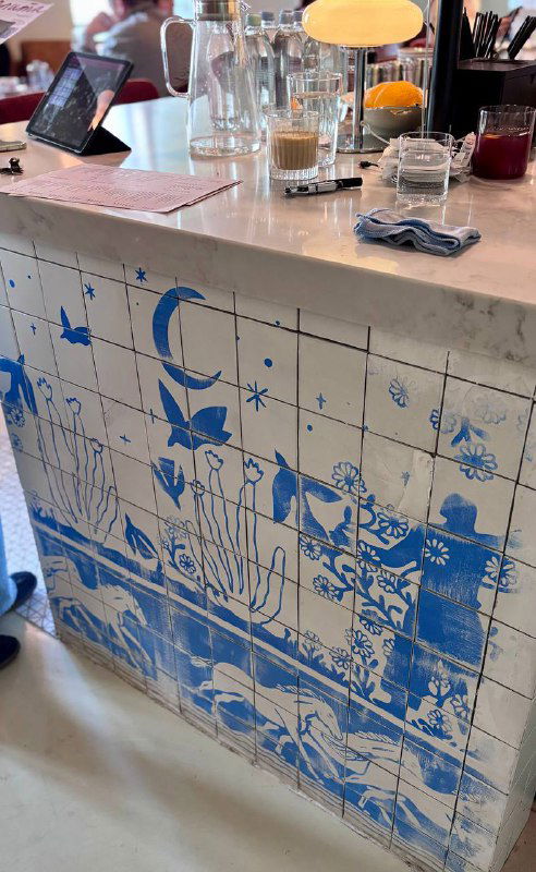 Мраморная стойка бара с керамическими синими плитками, стаканы и бутылки на фоне окна, декоративная посуда на прилавке.