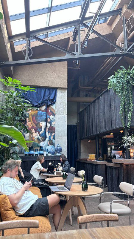 Интерьер кафе с высокими потолками и стеклянной крышей: столы, зелёные растения и гости, ощущение веранды внутри помещения.