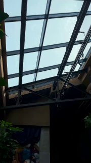 Фрагмент стеклянной крыши и металлических ферм над залом кафе: видно небо через стекло, конструкции и элементы декора интерьера.