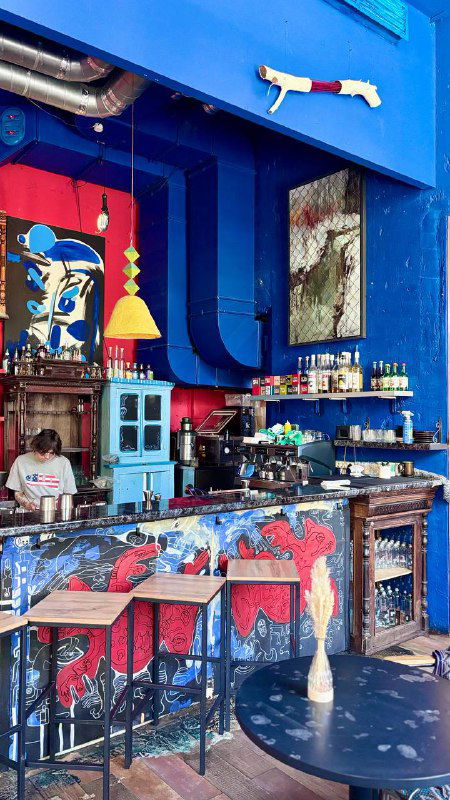 Барная зона на синем фоне: расписная стойка, бутылки на полках и декор, передача атмосферного эстетичного интерьера галерейного кафе в Москве.