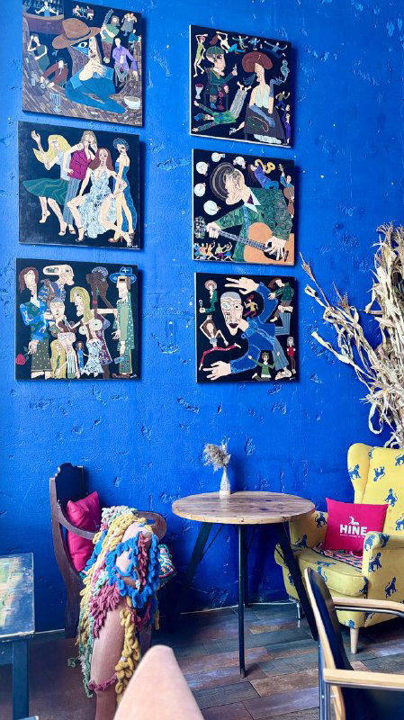 Голубая стена с развешанными картинами современных художников, небольшой столик у стены и декоративные элементы, интерьер галерейного кафе в Москве.