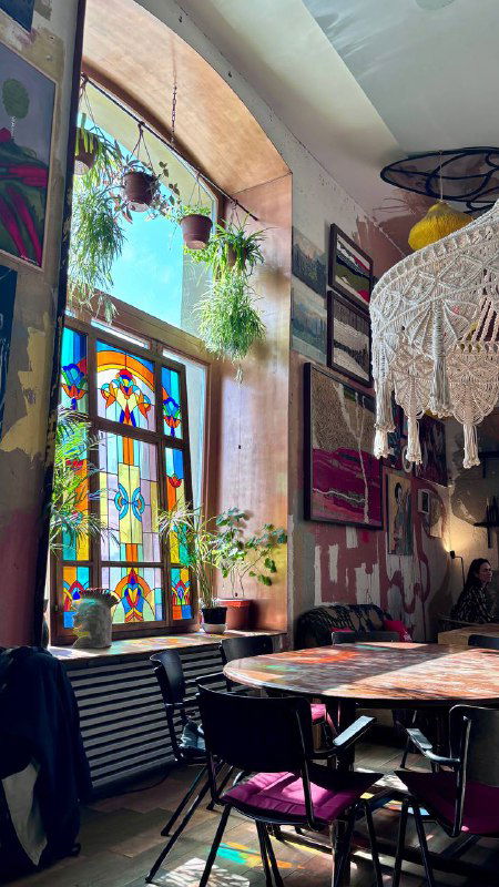 Большое витражное окно в зале ресторана, столы у окна, зелёные растения и солнечные блики на мебели — уютная обеденная зона в «Узоры», Москва.
