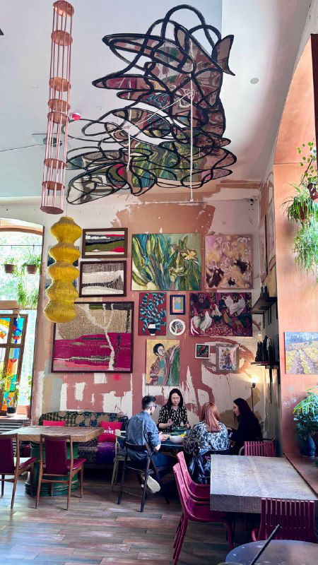 Высокий светлый интерьер с розовыми и синими акцентами, подвесные инсталляции и большая стена с арт-объектами — атмосфера творческого кафе в Москве.