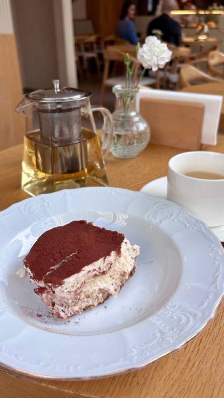 Кусочек тирамису на белой тарелке рядом с чайником и чашкой; десерт снят крупно на столе в уютном интерьере кафе Nobo в Москве.