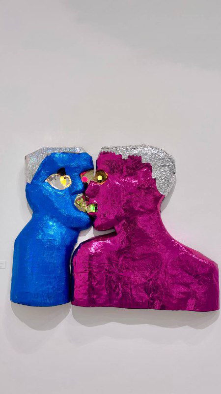 Скульптурная или настенная фигура в виде двух голов в ярких металлизированных синих и розовых тонах на белой стене музейного зала.