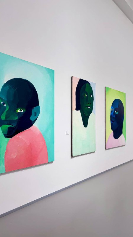 Ряд портретов на светлой стене: три картины с крупными стилизованными лицами в зелёно-розовой палитре, расставленные в линию.