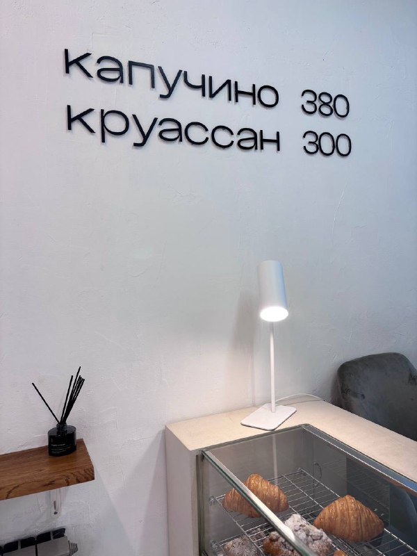 Интерьер кофейни с минимальным меню на стене: надпись «капучино 380 круассан 300», рядом лампа и витрина с выпечкой