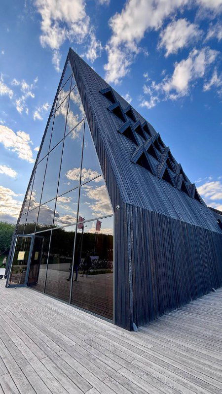 Угловое тёмное здание-центр посетителей с деревянной платформой и отражением неба в стеклах, современная архитектура.
