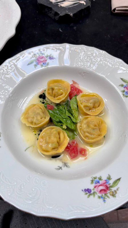 Тарелка с равиоли в светлом соусе, зелень и кусочки томата на фарфоровой посуде с цветочным узором, вид сверху.