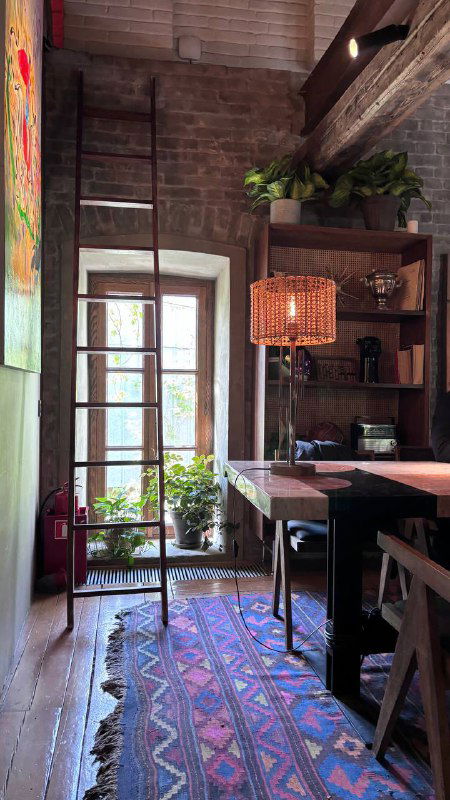 Уютный уголок с высоким окном, этническим ковром, деревянными полками и мягким светом — камерная зона для отдыха внутри заведения.