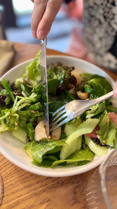 Процесс нарезки салата вилкой и ножом в тарелке с зеленью и кусочками овощей на деревянном столе ресторана.