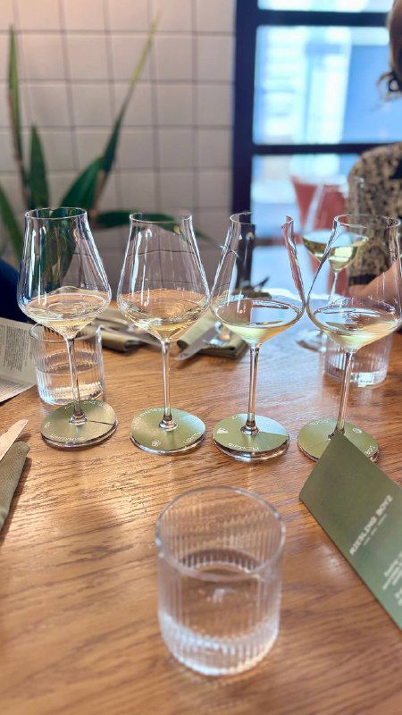 Дегустационные бокалы с белым вином на столе, подставки с логотипом и пустой стакан для воды; ресторанная атмосфера.