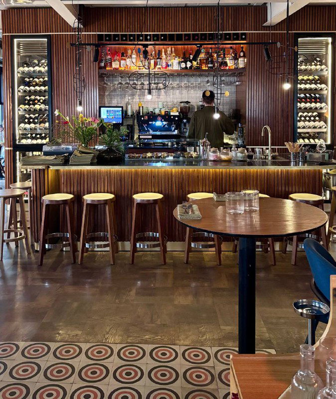 Барная стойка с витриной бутылок, стойками для барных стульев и деревянными столиками в переднем плане ресторана.