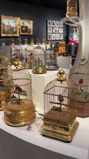 Несколько декоративных музыкальных шкатулок и автоматических клеток с фигурками птиц в светлой витрине, интерьер выставочного зала музея.