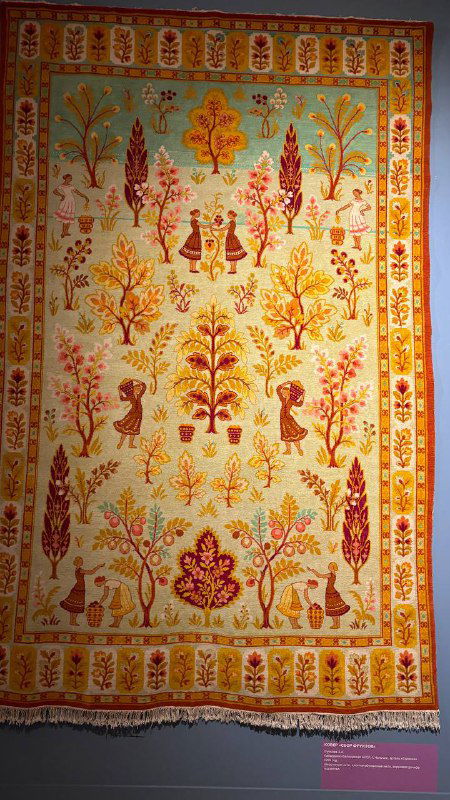 Большой светлый ковер с растительными мотивами и птицами, оформленный в традиционной манере и висящий на стене выставочного пространства.