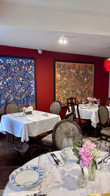 Уютный ресторанный зал с картинами и гобеленами на стенах, столы сервированы белыми скатертями и гжельской посудой.