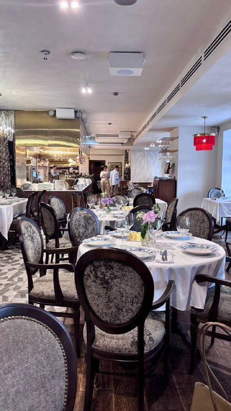 Ряд столов в ресторане с круглыми стульями и аккуратной сервировкой, в глубине видно открытую кухню и светлые потолки.