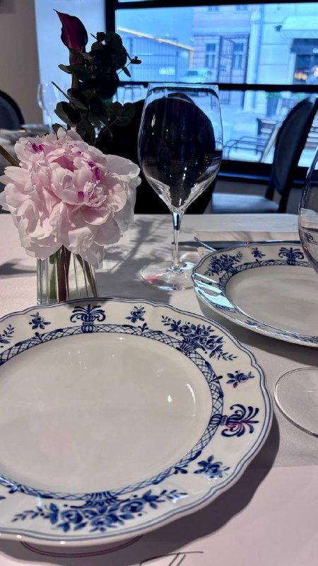 Стол у окна: гжельская тарелка, стеклянный бокал и нежно-розовая пионовидная композиция в небольшой вазе — близкий план.