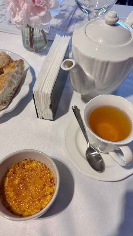 Чайник, чашка с чаем и крем-брюле на белой скатерти рядом с хлебной корзиной — десертная часть ужина на фоне сервировки.