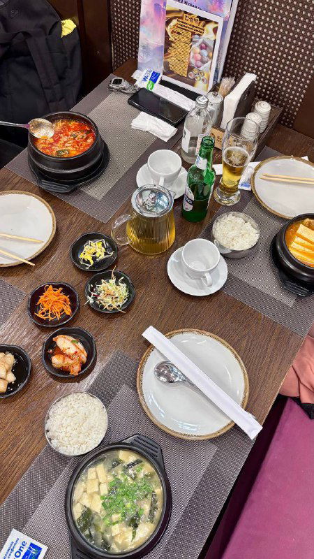 Стол с поданными корейскими блюдами: горячий суп в чугунке, маленькие закуски (бенчханы), чашки и бутылки напитков рядом на сервировке.