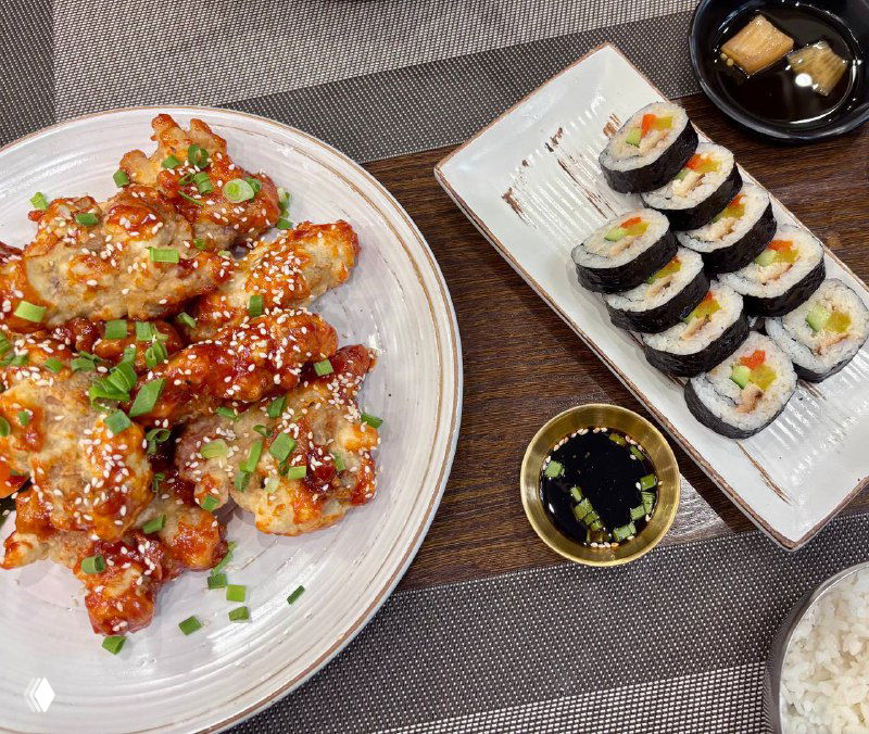 Крупный план блюда: глазированные острые куриные крылышки, рядом роллы кимпаб и соус — подача в простом, но аппетитном стиле.