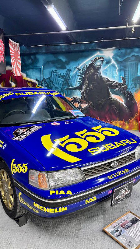 Синий гоночный Subaru с яркими жёлтыми эмблемами и цифрами на капоте, экспозиция в музее японских ретро-автомобилей, стенд с иллюстрациями позади.