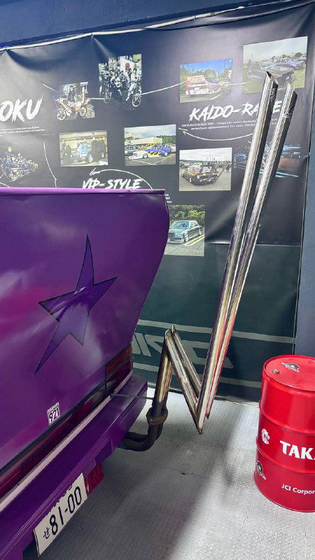 Крупный план задней части сиреневой машины: удлинённая выхлопная труба и элементы тюнинга, рядом стенды с описанием стиля и фотографиями.