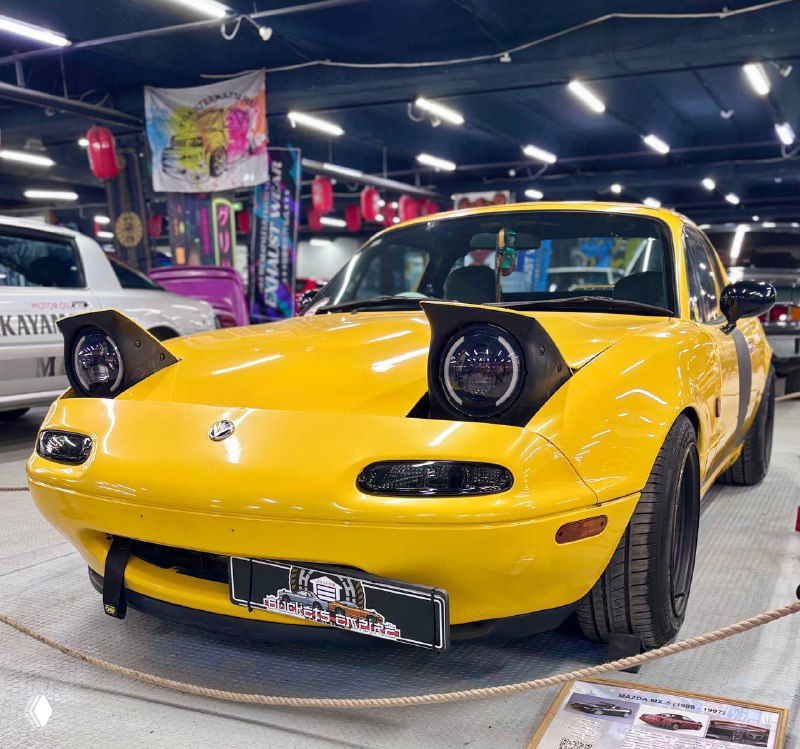 Жёлтый ретро-родстер с поднятыми фарами и мягкими формами кузова, выставлен на подиуме музея японских ретро-автомобилей среди других экспонатов.