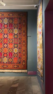 Большой геометрический ковер, висящий в музейном зале; тёплые коричневые и бордовые тона, крупный орнамент и аккуратная кайма по краю.