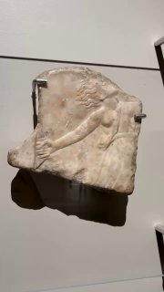 Фрагмент мраморного рельефа I века н.э.: частично сохранившаяся фигура в профиль, установленная на музейной стене с подсветкой.