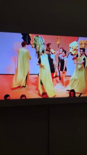 Кадр с экрана: группа артистов на сцене в жёлтых платьях, видеофрагмент перформанса, показанный в зале выставки.