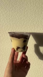 Стаканчик тирамису, снятый сбоку на светлом фоне; видна тёмная шоколадная прослойка и тень руки, минималистичная подача десерта.