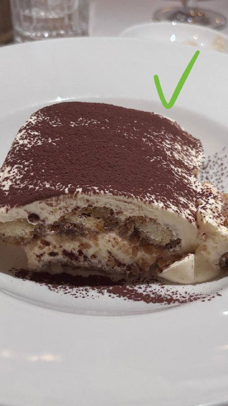 Кусок тирамису на белой тарелке крупным планом, сверху какао-порошок, видно слои крема и пропитанных коржей в разрезе.