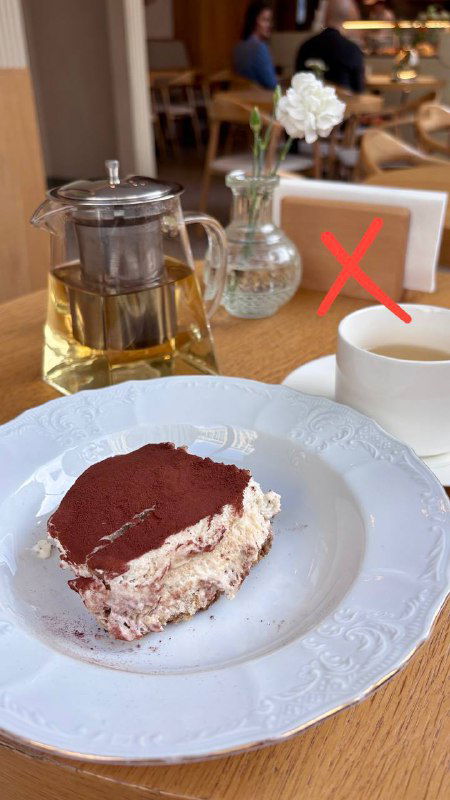 Кусок тирамису на тарелке в кафе рядом с чайником и чашкой; на кадре красный крест, обозначающий неудачную версию десерта.