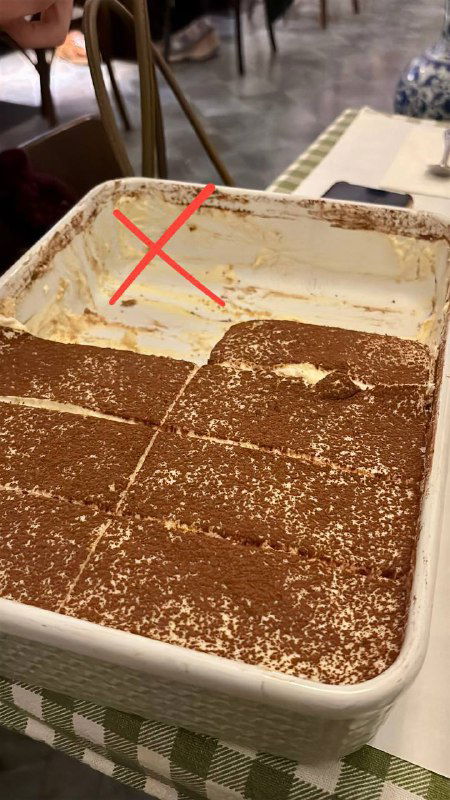 Большой противень тирамису с вырезанными порциями; видно остатки и отметку крестом, иллюстрирующее неудачный исход десерта.