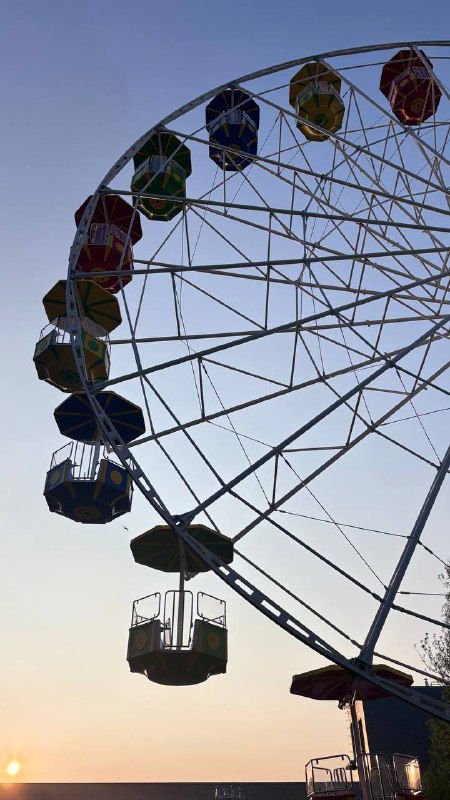 Крупное колесо обозрения на закате неба, силуэты кабинок и солнце низко над горизонтом — городской отдых в карельском лагере.