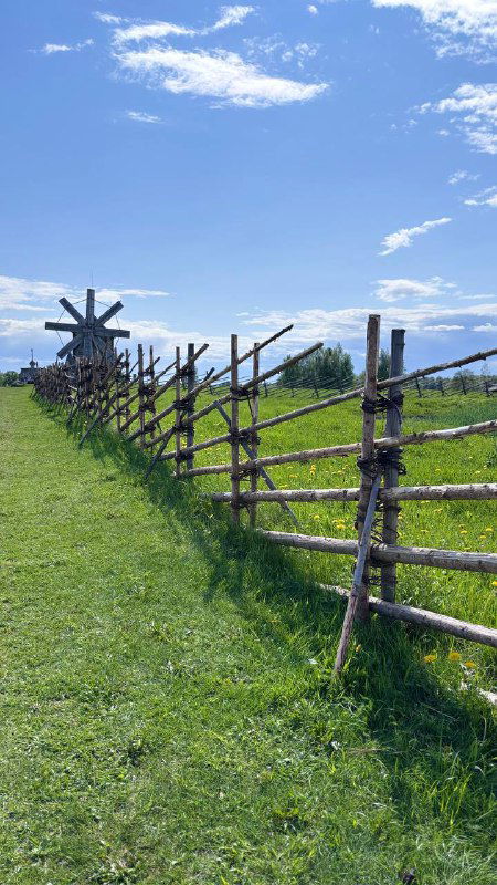 Традиционный деревянный забор и пологий луг с плотиной вдалеке, небо с облаками и тихая сельская сцена в Карелии.