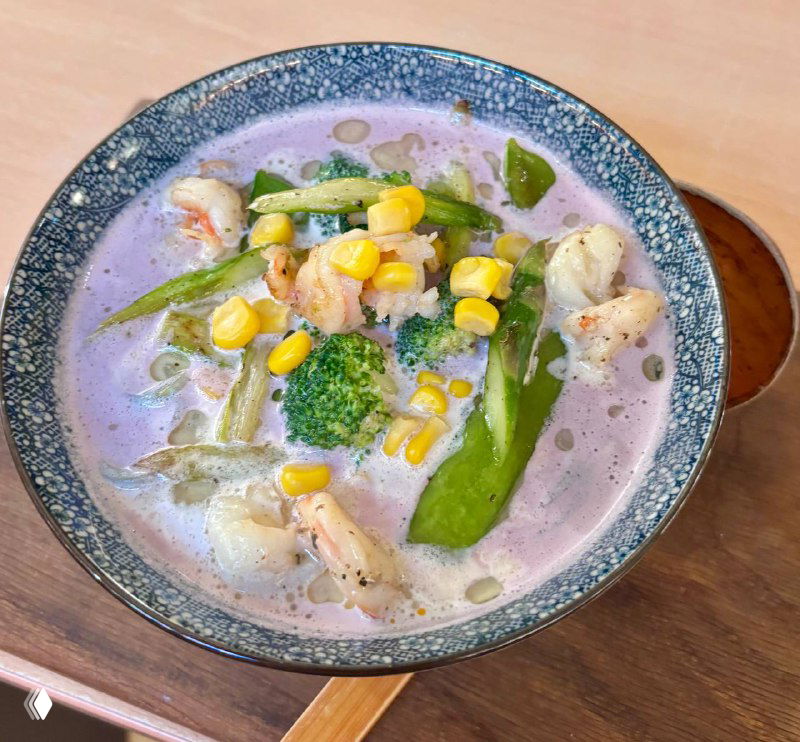 Миска фиолетового рамена с кусочками курицы, брокколи, кукурузой и зелёными стручками на деревянной подставке — специальный рамен с фиолетовым бататом.