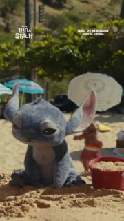 Игрушечный Ститч, оставленный в песке на пляже: фигурка в песочном окружении, зонтики и ведёрко на заднем плане, кадр из трейлера или тизера.