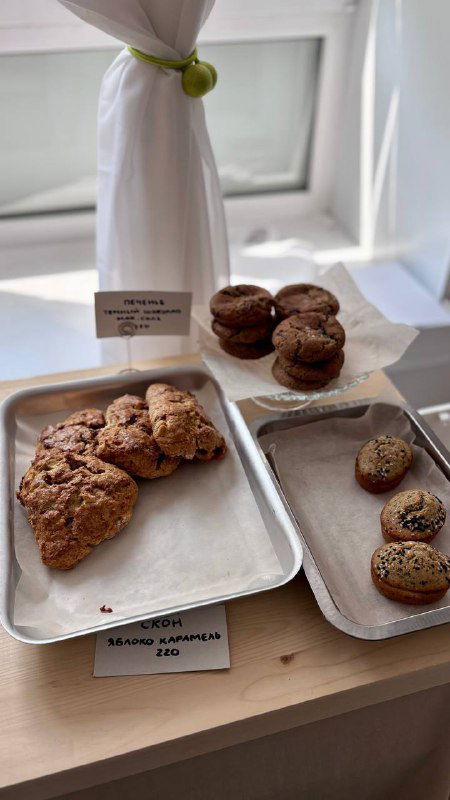 Подносы с ассортиментом печенья, мини‑пирожными и булочками у окна: коричневые печенья и небольшие пирожные на подносах рядом с ценниками