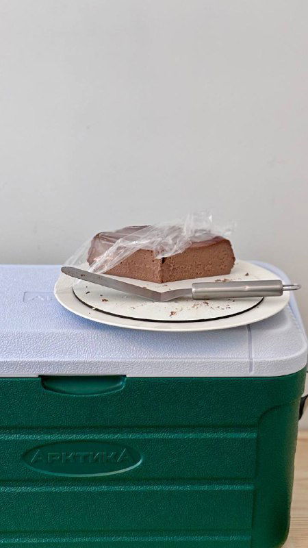 Кусок шоколадного сан‑себастьяна на бумажной тарелке и нож, торт плотной структуры стоит на изотермическом контейнере — демонстрация текстуры десерта