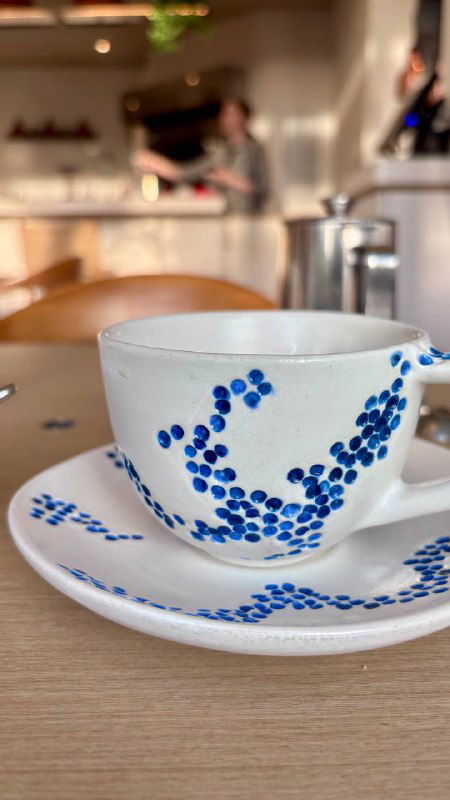 Крупный план керамической чашки с синим узором на блюдце, на заднем плане стол и интерьер ресторана Claudia — акцент на посуде бренда.