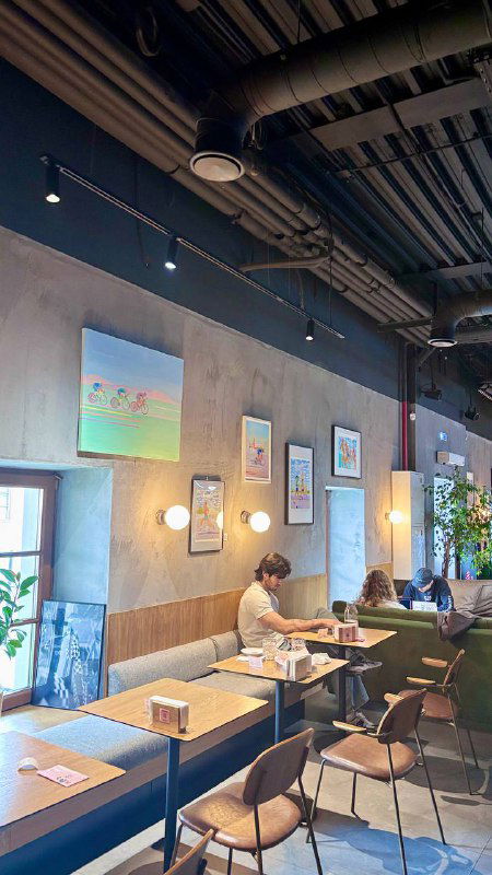 Еще один угол интерьера с настенными картинами и столиками, посетитель за ноутбуком; пространство подходит для встреч и работы вне дома.