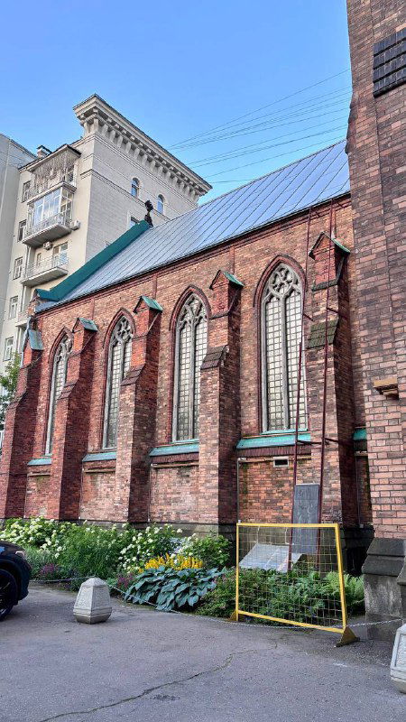 Боковой фасад англиканской церкви в окружении городских домов и небольшого сквера; кирпичная стена с узкими готическими окнами и цветниками.